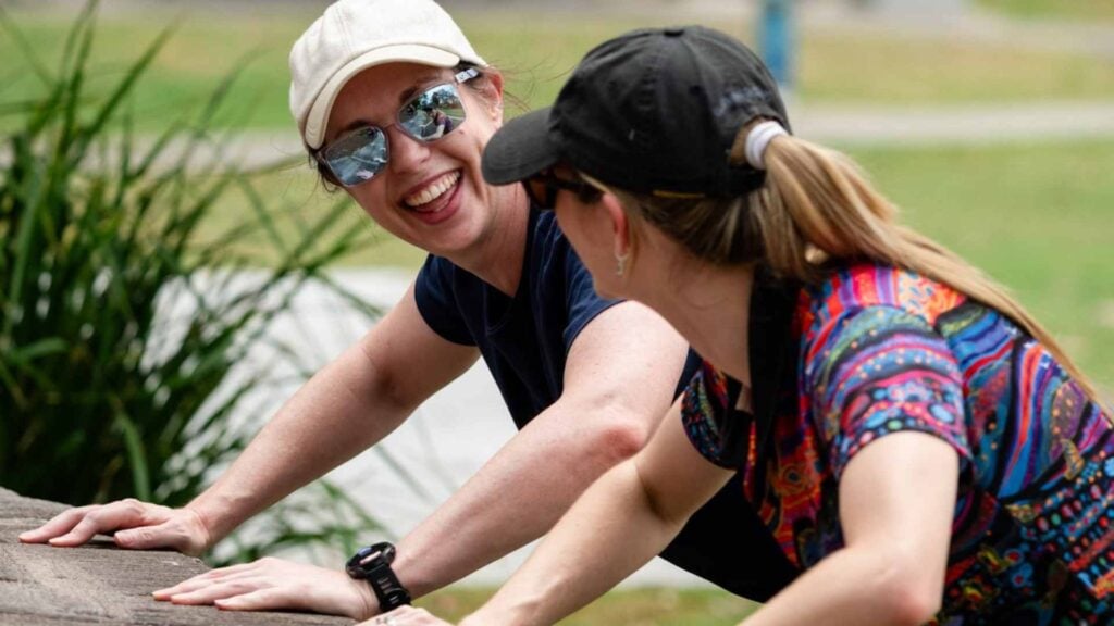Two women do modified push ups on stone wall in public space