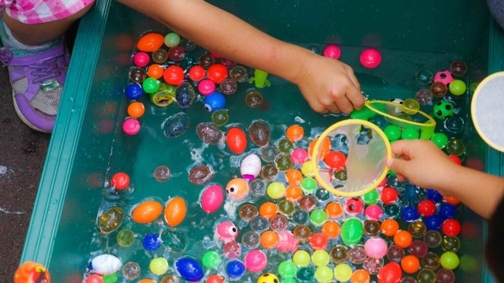 Young children engage in water play, scooping colourful balls into a net from a play tub