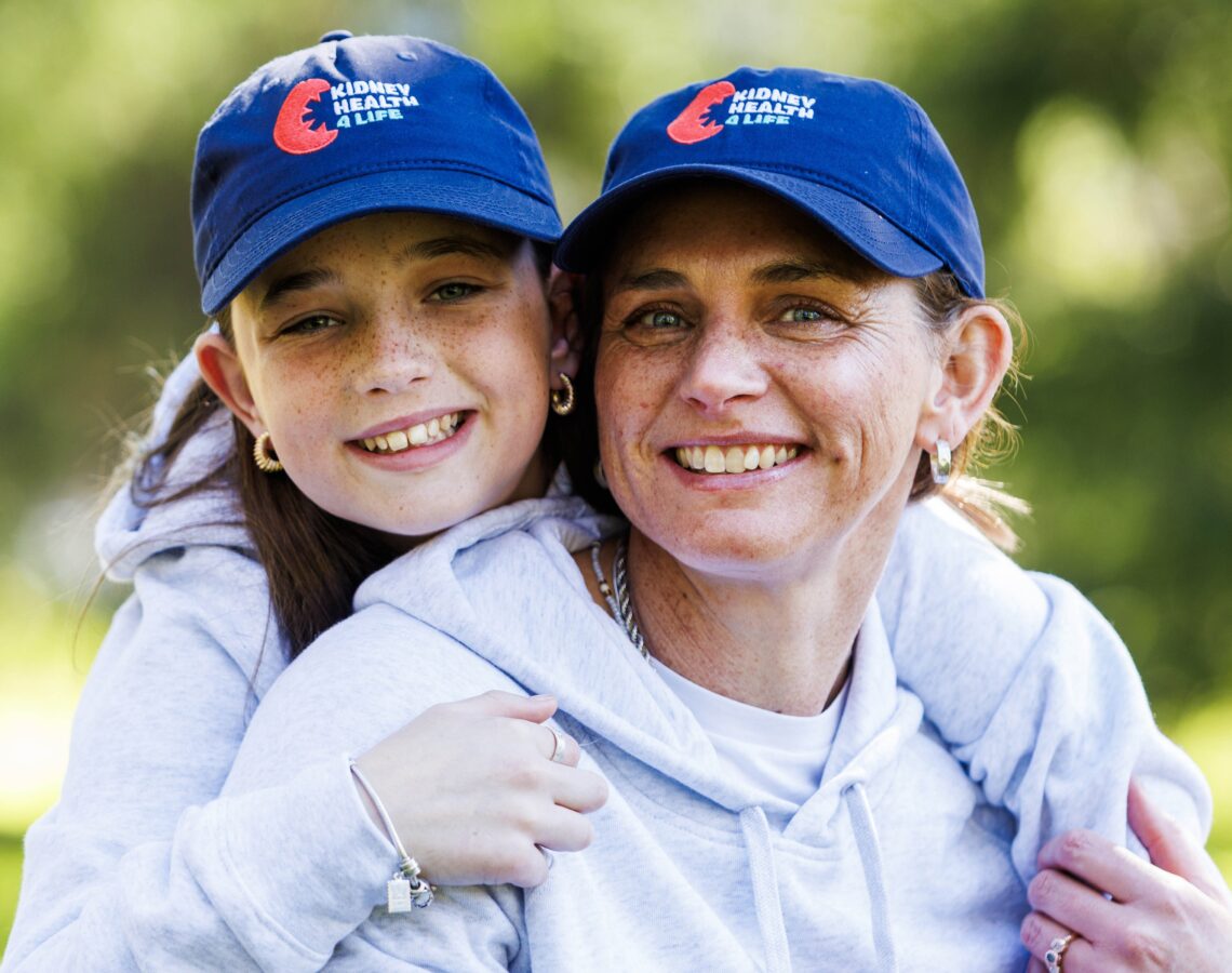 Young girl hugging woman, both in grey hooded sweatshirts, wearing blue 'Kidney Health 4 Life' hats.
