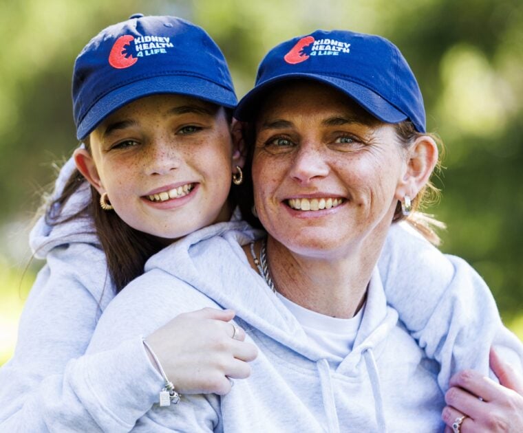 Young girl hugging woman, both in grey hooded sweatshirts, wearing blue 'Kidney Health 4 Life' hats.