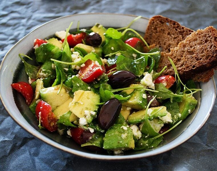 Bowl which features salad leaves, tomato, olives, avocado and two wholegrain crackers.