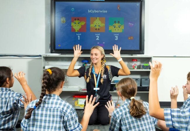 Woman sits on the ground surrounded by primary school children teaching about cybersecurity
