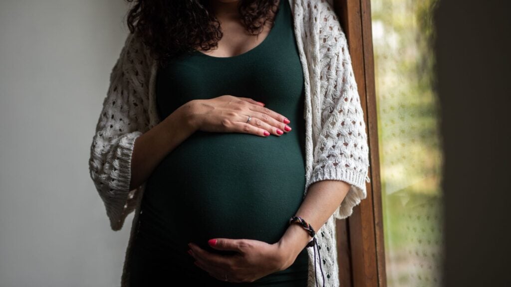 Woman in green dress holds her baby bump standing beside a window
