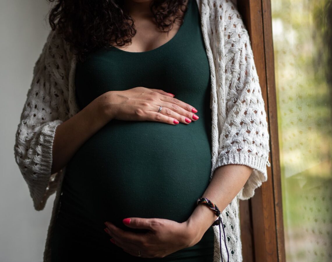 Woman in green dress holds her baby bump standing beside a window