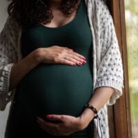 Woman in green dress holds her baby bump standing beside a window