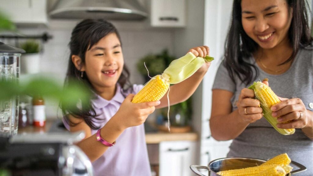 Mother and daughter preparing corn cobs and vegetables in the kitchen