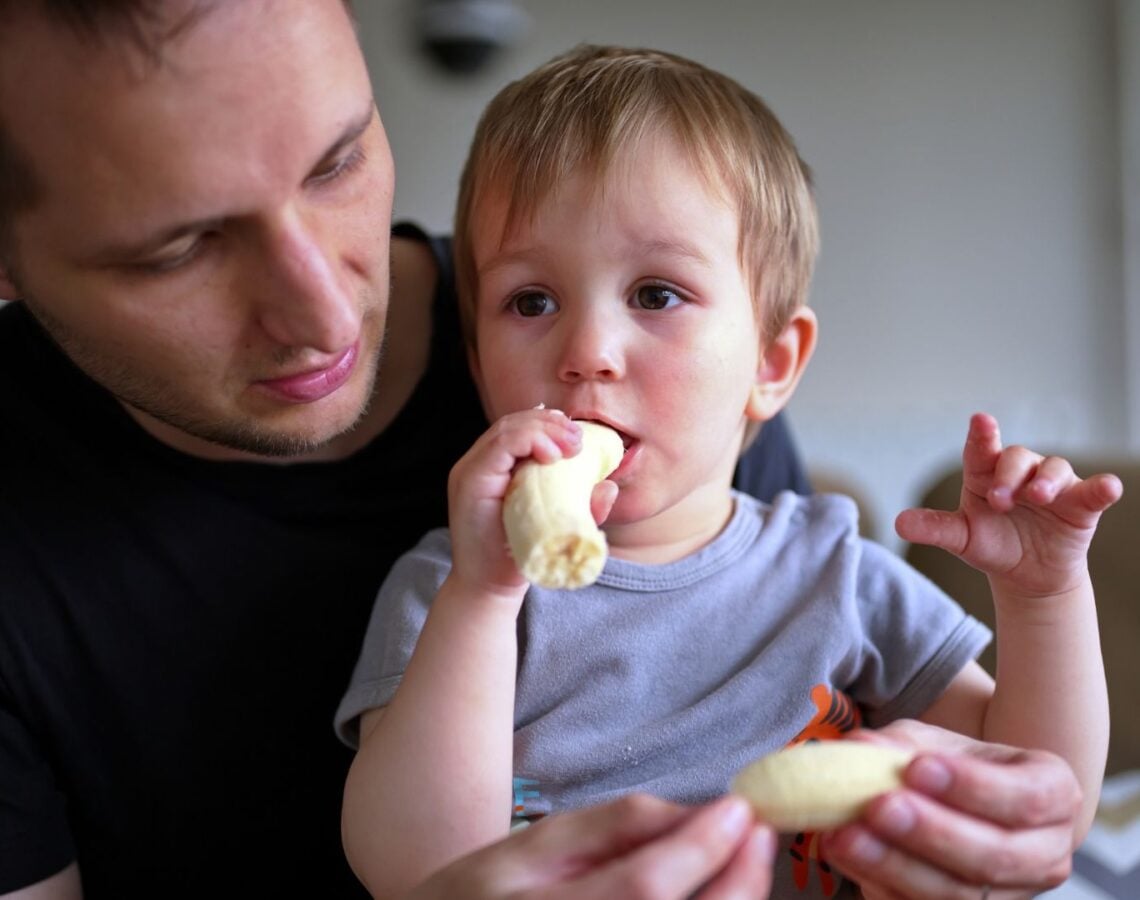 Young boy eats a banana while sitting in his father's lap