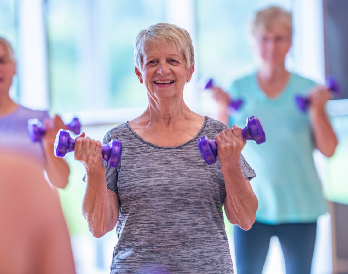 Woman standing in a exercises class holding two purple weights.