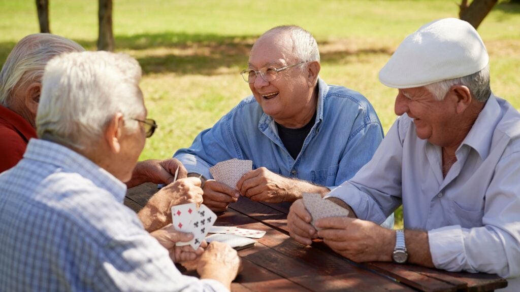 4 older men sit at a table outside playing cards