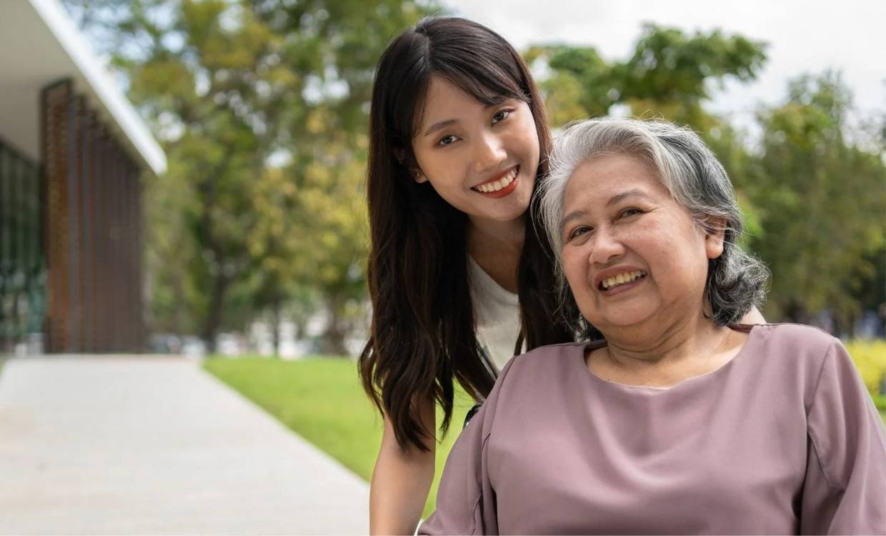 A young woman supports an older adult using a wheelchair while walking together along an outdoor path