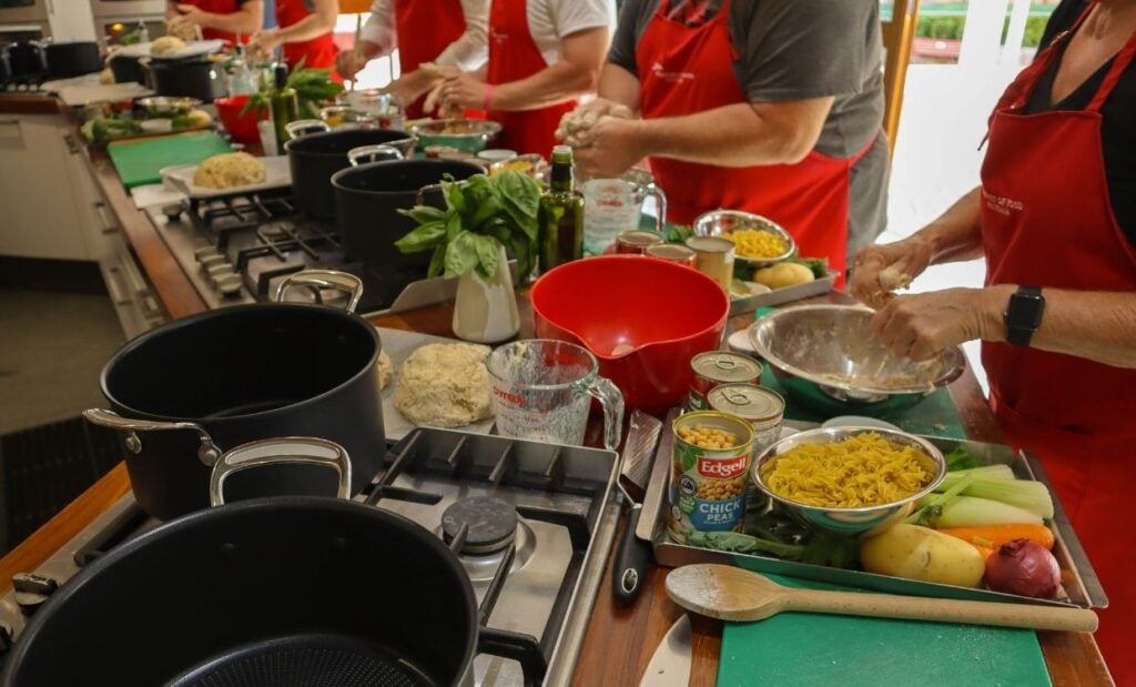 Cooking class with participants standing at kitchen workbenches, preparing ingredients and using a variety of cooking utensils