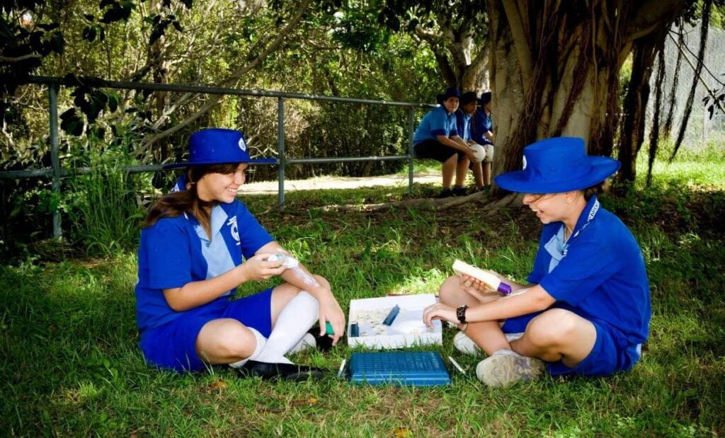 Two schoolgirls wearing sun-safe clothing and applying sunscreen while enjoying an outdoor game together