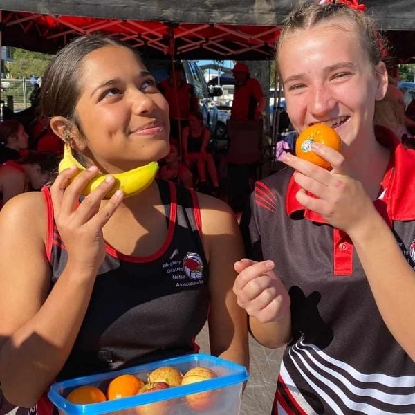 Two teenage girls enjoy fresh fruit together in front of a team marquee at a sports carnival