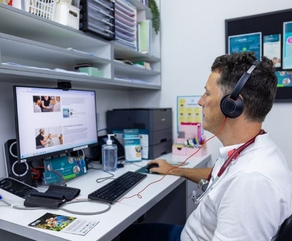 Health professional GP sitting at a workspace in a consultation room, wearing a headset and listening to Clinician’s Guide Podcast series