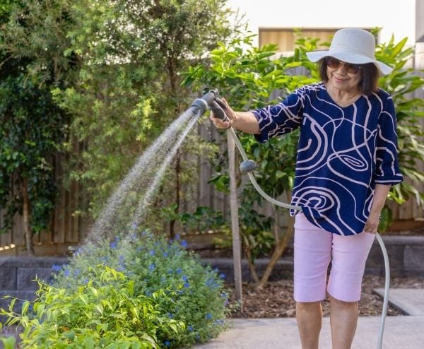 Older woman wearing sun safe clothing watering her outdoor garden with a hose